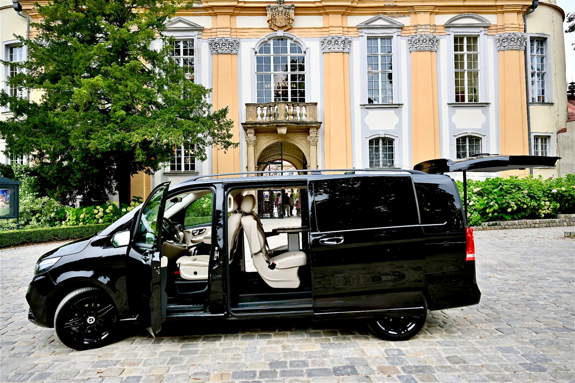 Czarny Mercedes V klasa z otwartymi elektrycznie drzwiami bocznymi i klapą bagażnika przed pałacem
